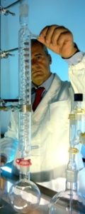 Il dottor Lorenzo Domini con il camice e cravatta rossa dietro una serpentina in vetro borosilicato connessa ad un pallone evaporatore mentre lavora in laboratorio ad un distillatore verticale in vetro