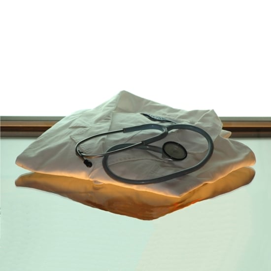 Piano di vetro trasparente con un camice da medico pulito e piegato appoggiato sopra il piano con uno stetoscopio; sfondo luminoso bianco