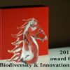Immagine del premio ricevuto dalla azienda Casalvento rappresentato da un cavallo in cristallo trasparente su sfondo rosso e nero con riflessi su vetro e scritta nera: premio per biodiversità ed innovazione