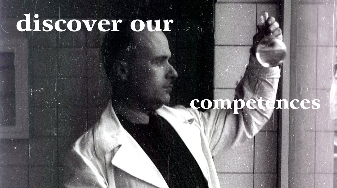 Immagine in bianco e nero del professor Giovanni Domini con il camice e maglione scuro mentre controlla la qualità di un estratto contenuto in un pallone di vetro da laboratorio alla finestra sullo sfondo di mattonelle di ceramica