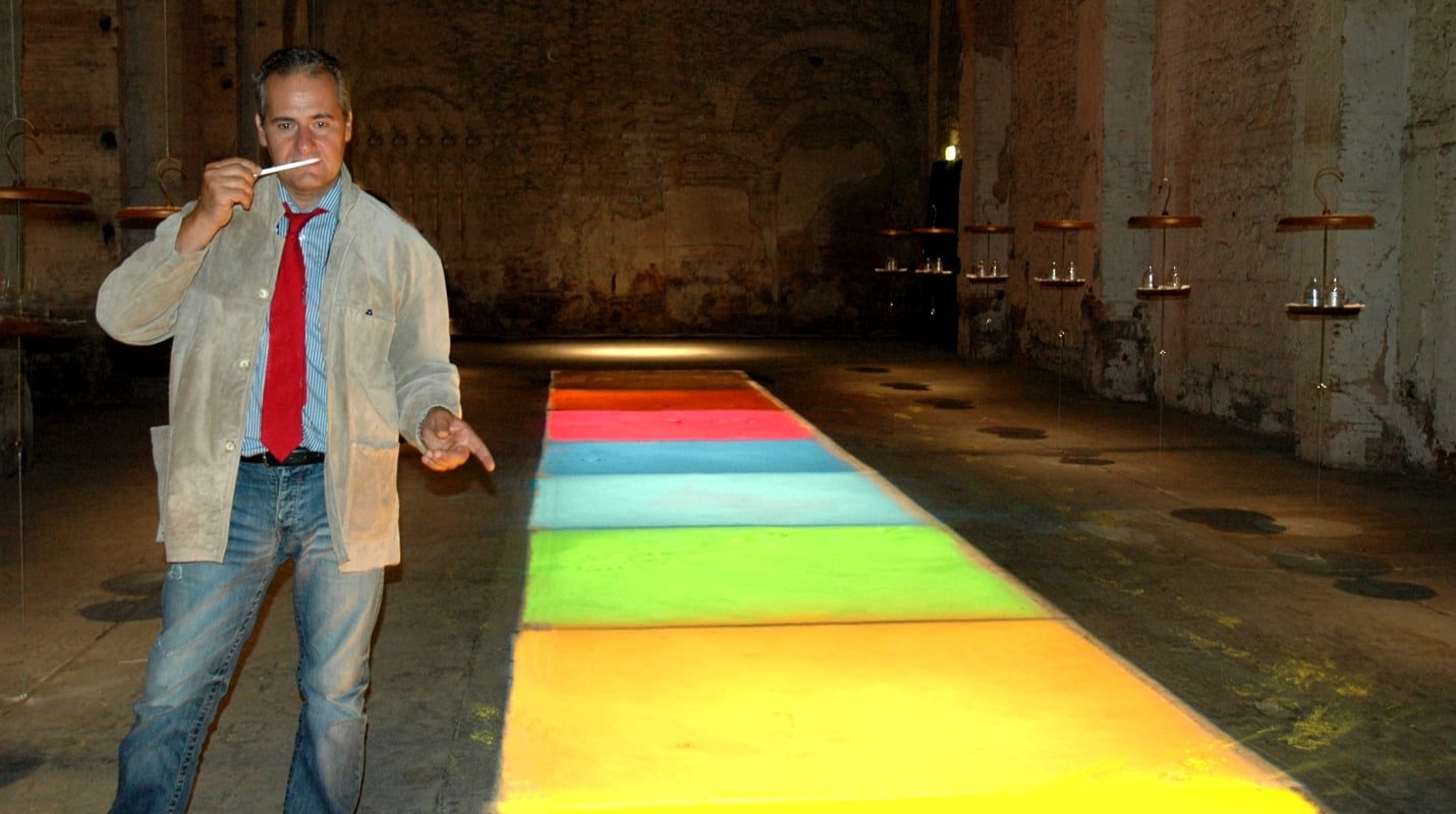 Luminous panels with the colors of the rainbow like a luminous street, image of Lorenzo Domini in jeans, striped shirt and red tie while smelling a perfumed paper on a dark rustic background