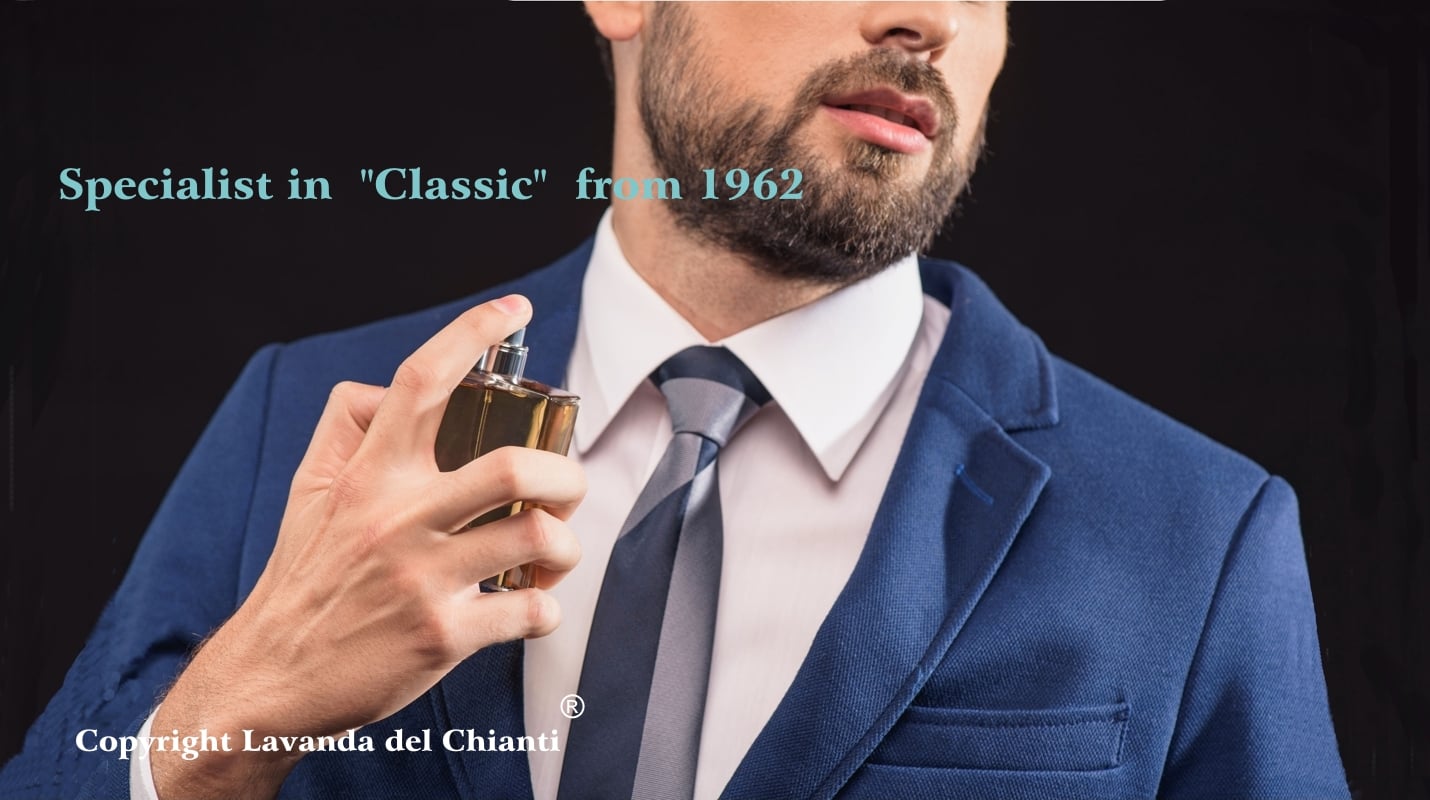 Young bearded man wearing blue jacket and gray tie over pink shirt spraying some perfume on his neck against black background; blue lettering: specialists in classics since 1962 and white lettering: property of Lavanda del Chianti